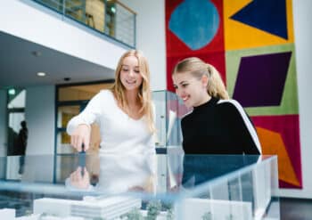 Zwei junge Frauen betrachten gemeinsam ein Architekturmodell der Zentrale von Deichmann. Im Hintergrund ist eine farbenfrohe, geometrische Wanddekoration in einem modernen Gebäude sichtbar.