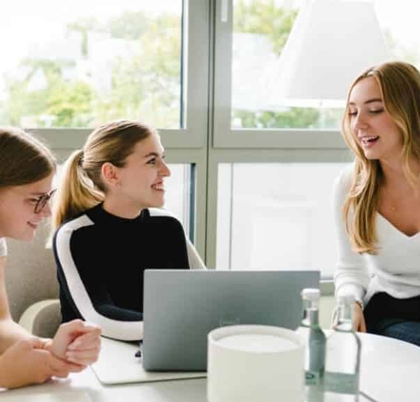 Drei Frauen unterhalten sich lächelnd um einen Tisch. Eine hat einen Laptop vor sich. Im Hintergrund sind Fenster mit Tageslicht. Eine Flasche Wasser steht auf dem Tisch.