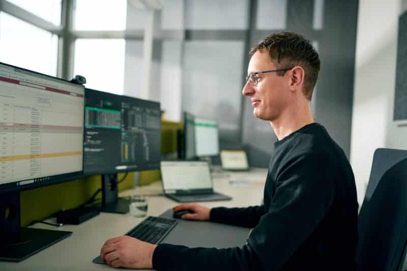 Ein Mann arbeitet am Computer im Büro und blickt auf zwei Bildschirme mit Tabellen und Code.