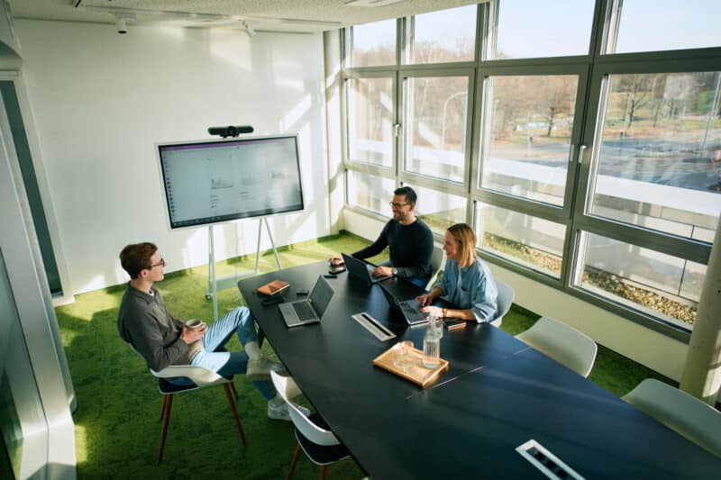 Drei Personen sitzen an einem Konferenztisch, arbeiten mit Spaß an Laptops. Ein großer Monitor zeigt Produktbilder. Heller Raum mit großen Fenstern und grünem Teppich.