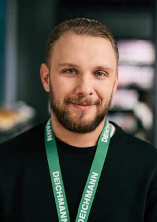 Ein junger Mann mit grünem "Deichmann"-Schlüsselband lächelt. Er trägt einen schwarzen Pullover.