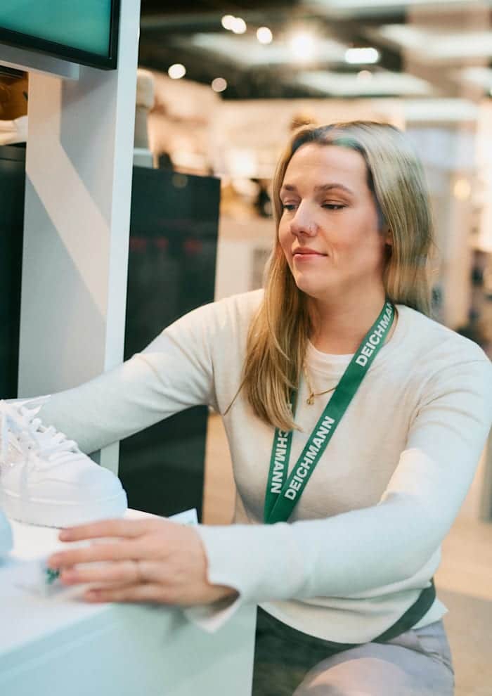 Eine Verkäuferin mit einem "Deichmann"-Schlüsselband arrangiert weiße Sneaker auf einem Regal in einem gut beleuchteten Schuhgeschäft.