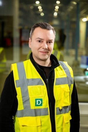 Person trägt eine gelbe Warnweste mit reflektierenden Streifen und einem grünen Aufnäher mit weißem Buchstaben D in einer Lagerhalle
