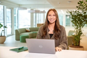 Frau mit langen braunen Haaren sitzt an einem weißen Tisch vor einem geöffneten silbernen-Laptop in einem modernen Büro mit Sofa und Pflanzen im Hintergrund.