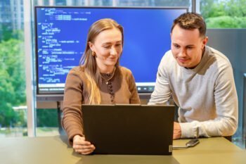 Zwei Personen sitzen an einem Tisch und schauen auf einen Laptop, im Hintergrund ein Bildschirm mit Programmiercode.