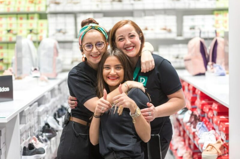 Drei lächelnde junge Frauen umarmen sich in einem Geschäft mit Schuhregalen im Hintergrund. Eine Frau zeigt Daumen hoch. Umgebung ist hell und geschäftig mit vielen ausgestellten Schuhen.