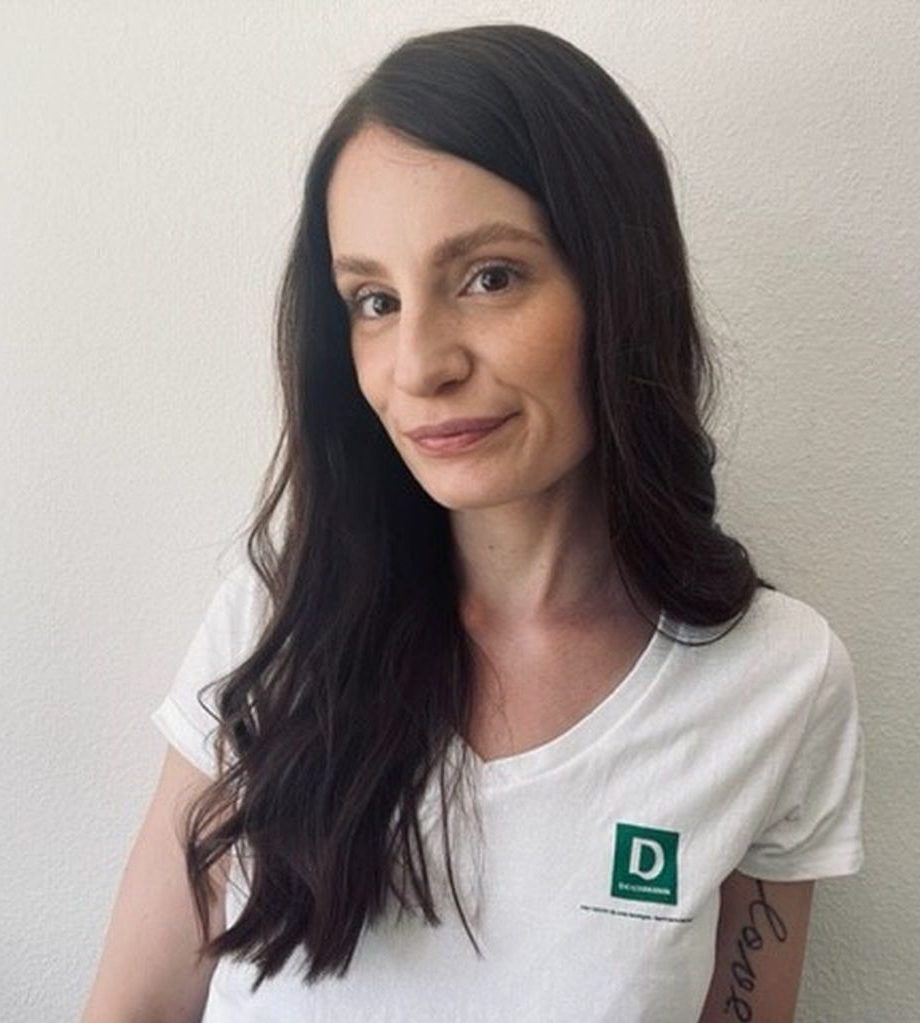 Eine Frau trägt ein weißes T-Shirt mit grünem "Deichmann"-Logo. Sie steht in einem neutralen Innenraum und blickt in die Kamera.