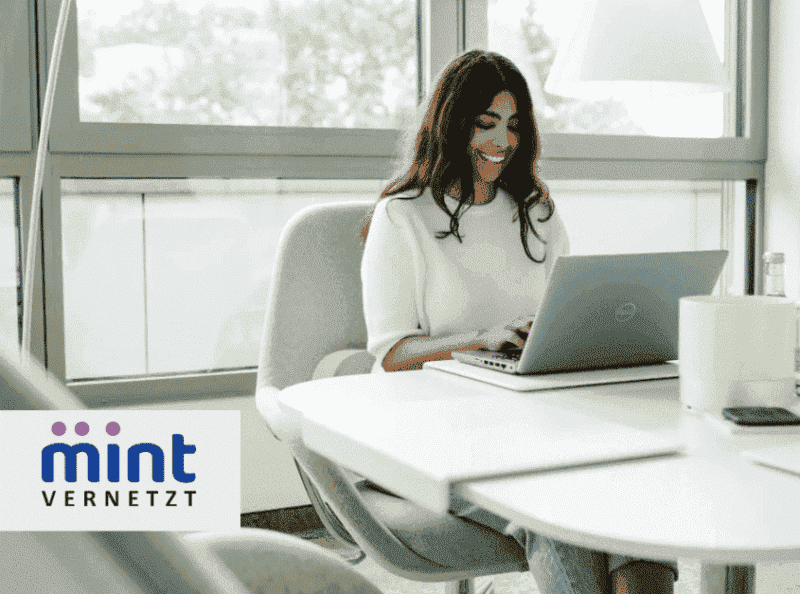 Junge Frau mit langen dunklen Haaren arbeitet lächelnd an einem Laptop in einem modernen Büro, unten links das Logo von MINTvernetzt.