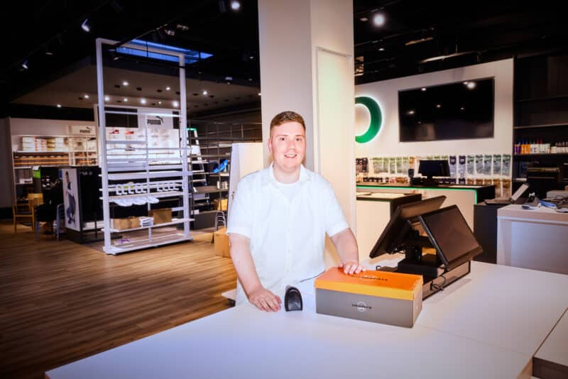 Ein junger Mann steht lächelnd an einem Verkaufstresen in einem modernen Schuhgeschäft. Vor der Person liegt ein Karton, umgeben von Regalen und einem großen Bildschirm im Hintergrund.