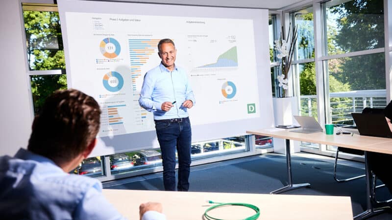 Ein Mann präsentiert vor einem großen Bildschirm mit Diagrammen und Statistiken, darunter Text "Phase 2: Aufgaben und Status". Im Hintergrund sind Fenster mit Blick nach draußen.