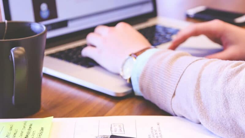 Eine Person tippt auf einem Laptop im Büro, umgeben von einem Kaffeebecher, Notizen und einem Smartphone.